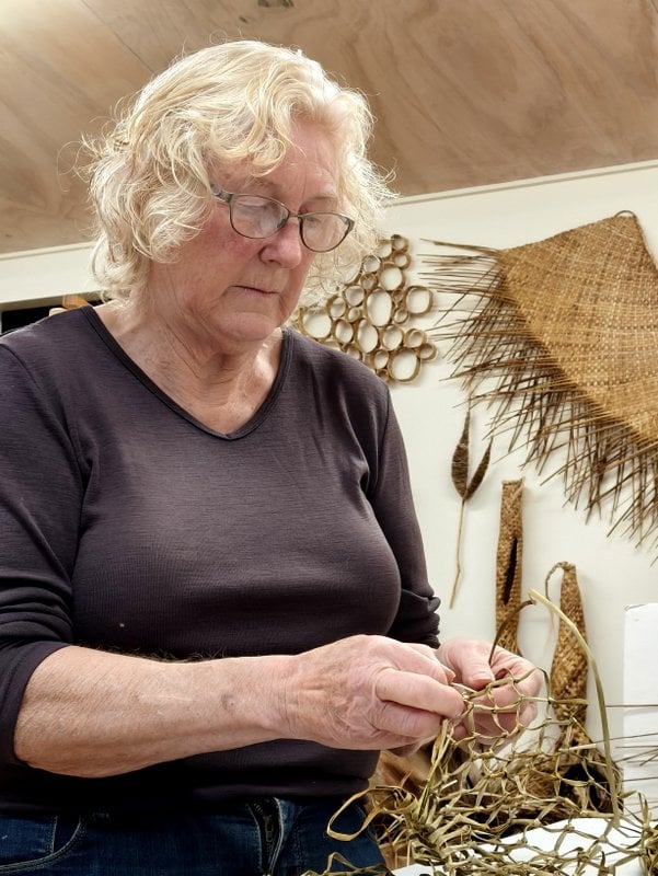 Anne Daniel creating with harakeke in her studio, Artworks in the background.